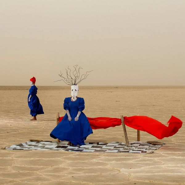 A woman in a blue dress sits on a red bench in the desert wearing a mask made from a jerry can. Another woman stands in the background.