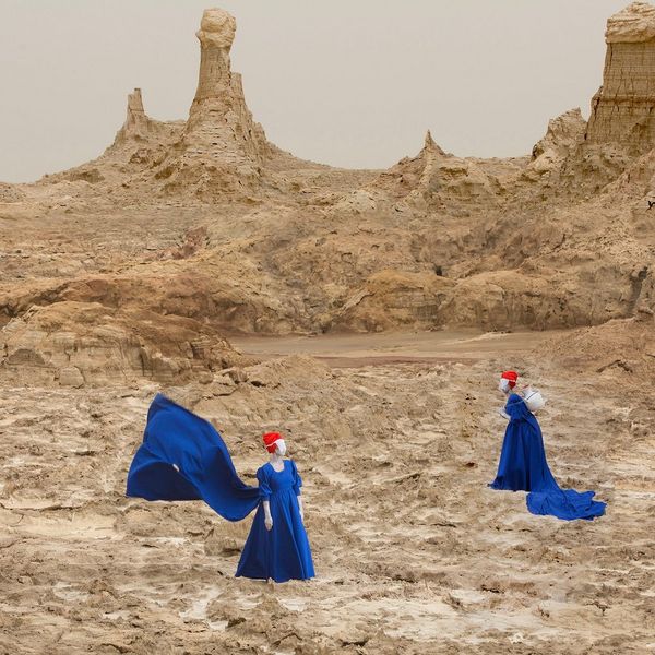 Two women in blue dresses with flowing trains stand in a desert landscape.