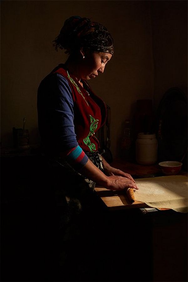 A Kazakh woman rolls out pastry with a wooden rolling pin in a darkened room with light coming from one side