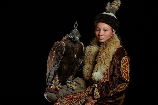 A young Kazakh boy wearing traditional clothing, with a fur collar and warm hat, sits with a hooded Golden Eagle on his arm