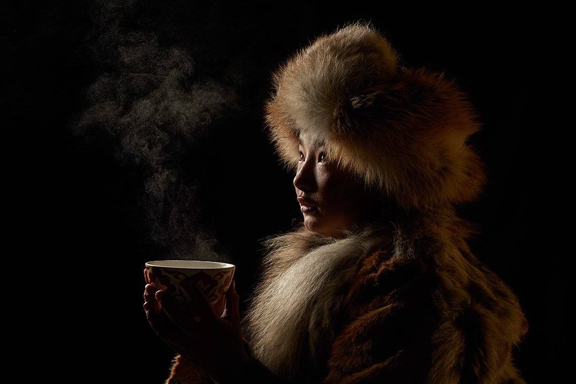 Dim light falls on the raised face of Damel, a 13-year-old Kazakh girl wrapped in a heavy fur coat and headdress, as she looks out of shot, with wisps of steam rising from the cup of tea she is holding in both hands.