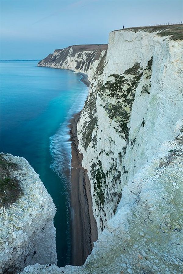 White cliffs seen from above stretching down to the beach. They look bluish-white and have a tiny figure on top in the distance. Photo by David Noton.