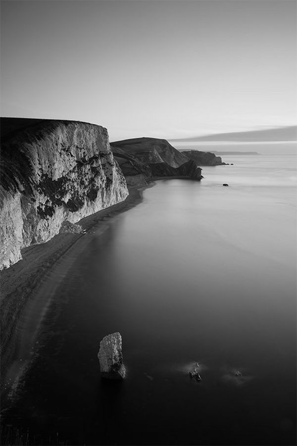A black and white beach landscape, with rock stacks in the sea and cliffs behind, photographed by Helen Bartlett.
