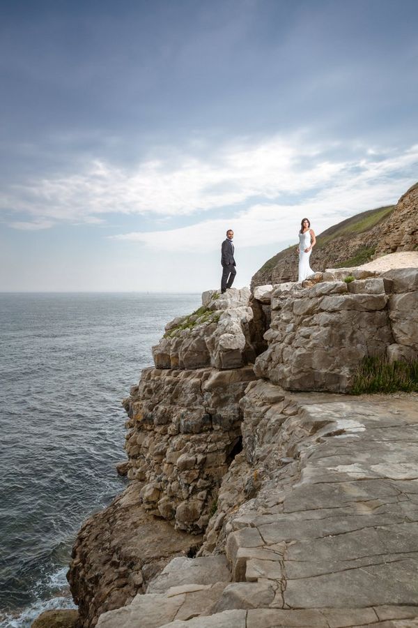 A bride and groom stand on a coastal path, photographed from below by Sanjay Jogia.
