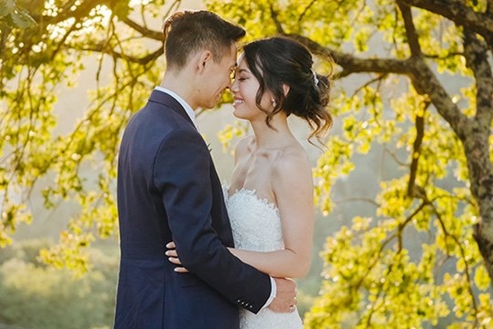 A bride and groom stand close together, faces touching, in front of a tree full of yellow blossoms.