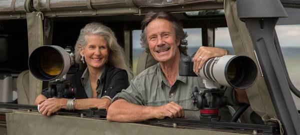 Photographers Jonathan and Angela Scott lean on the side of a camouflaged window, their Canon cameras with telephoto lenses set up in front of them, ready to photograph wildlife.