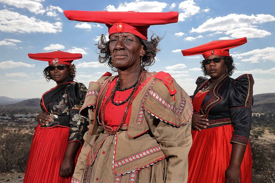 Three Herero women stand in a symmetrical triangular pose, wearing red, khaki and black colonial-style dresses and jackets. They also wear red wide-brimmed hats that lift up at each end like cows horns.