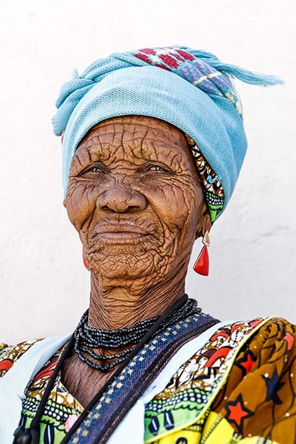 An elderly Namibian woman wearing a colourful patterned dress, dark blue beaded necklace, light blue headscarf, and earrings with large, triangle-shaped red stones.