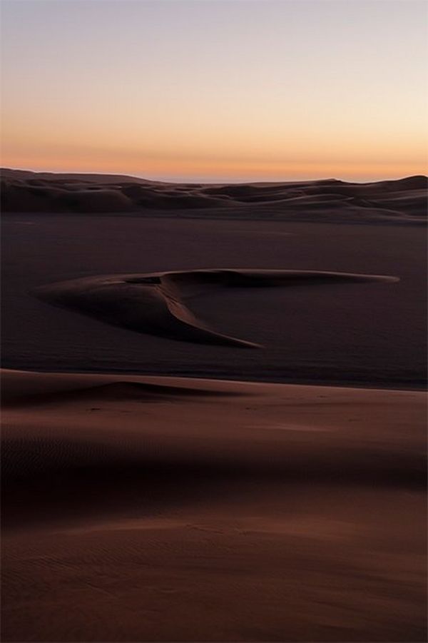 The dunes of the Namibian desert rise to undulating ridges in the distance, with a distinctive dune shaped like a barbed arrowhead in the middle distance.