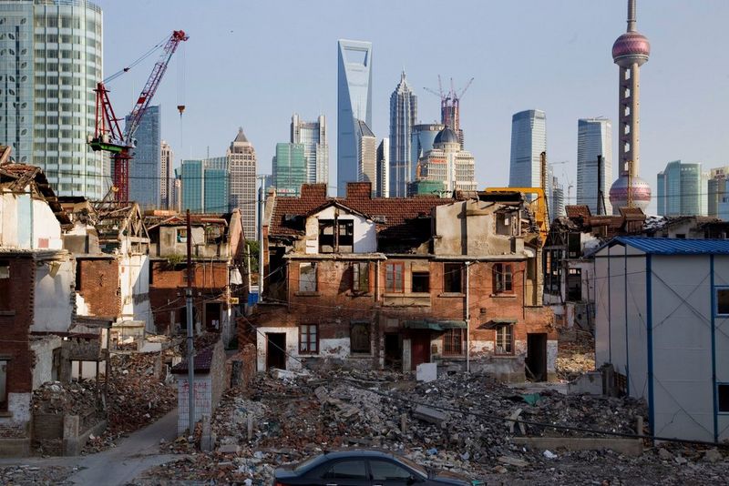 Old buildings being torn down, leaving piles of rubble in the foreground, with cranes and skyscrapers visible in the background. 