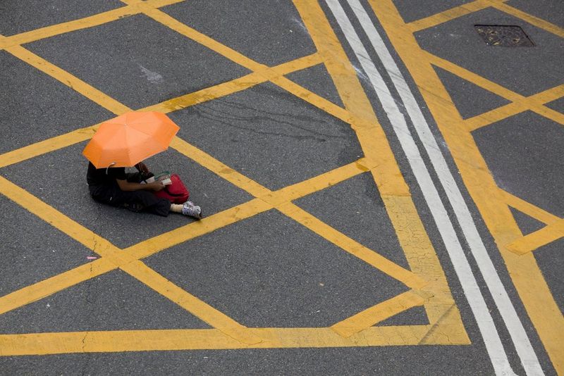 A person with an orange umbrella sitting on the tarmac of a yellow box junction.