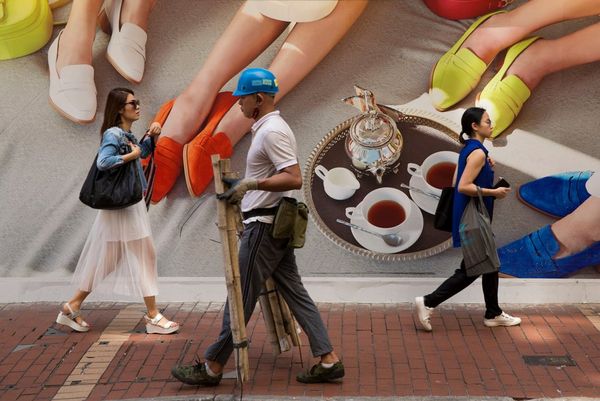 Three pedestrians walking along a Hong Kong street past a brightly coloured poster.