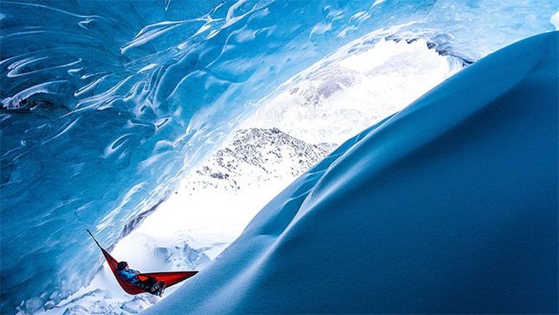 A man walks beside a still lake fringed with fir trees, under a low sun.