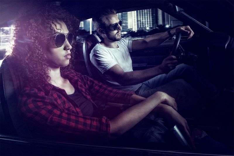 A man and woman are seen driving a car, sunlight streaming in from behind.