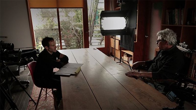 Bryan Fogel and Grigory Rodchenkov sit opposite each other at a long wooden table.