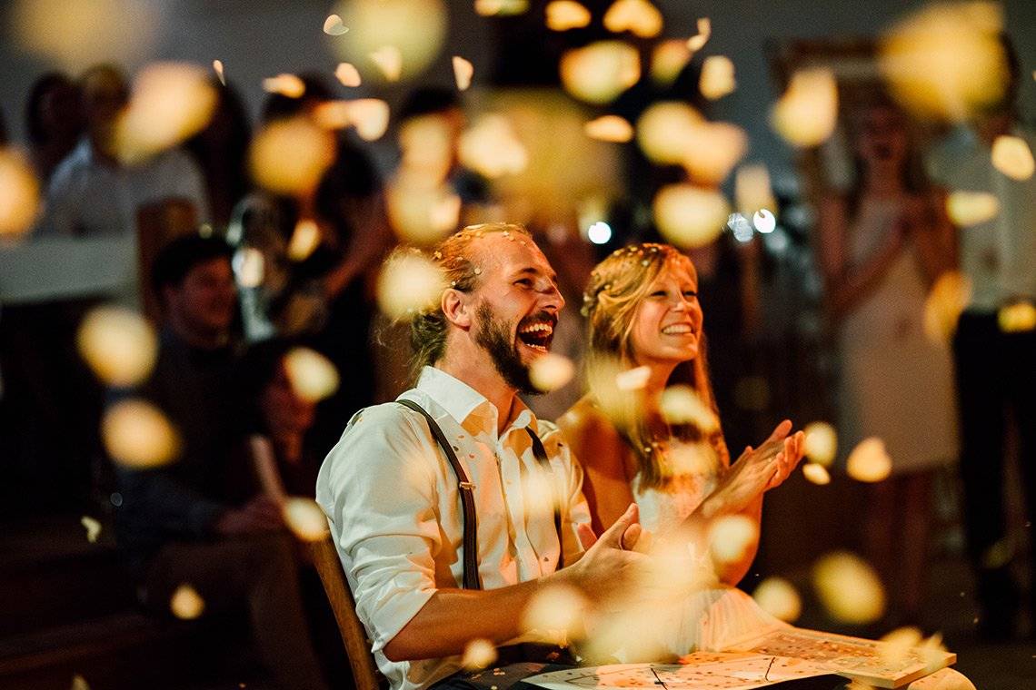 A couple laughing in delight as confetti falls around them. They are in sharp focus, everything else is blurred. Photo by Markus Morawetz.