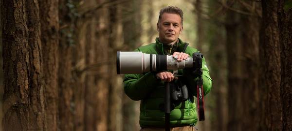 Chris Packham stands in a woodland wearing a green jacket, his hands resting on his Canon 5DS R fitted with a Canon telephoto lens, on a stand.