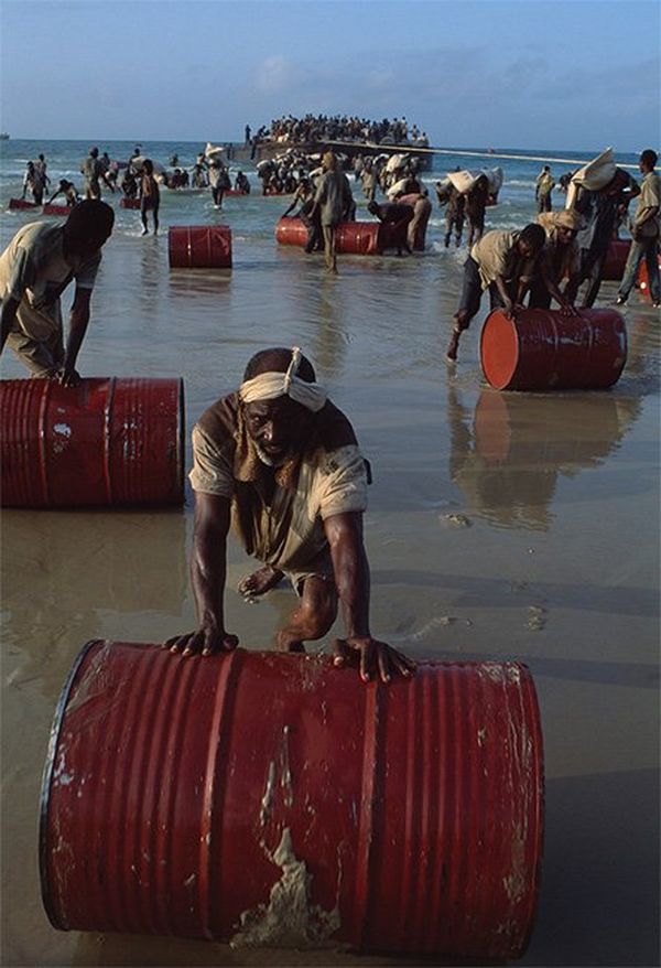 Men roll red barrels out of the sea onto the beach.