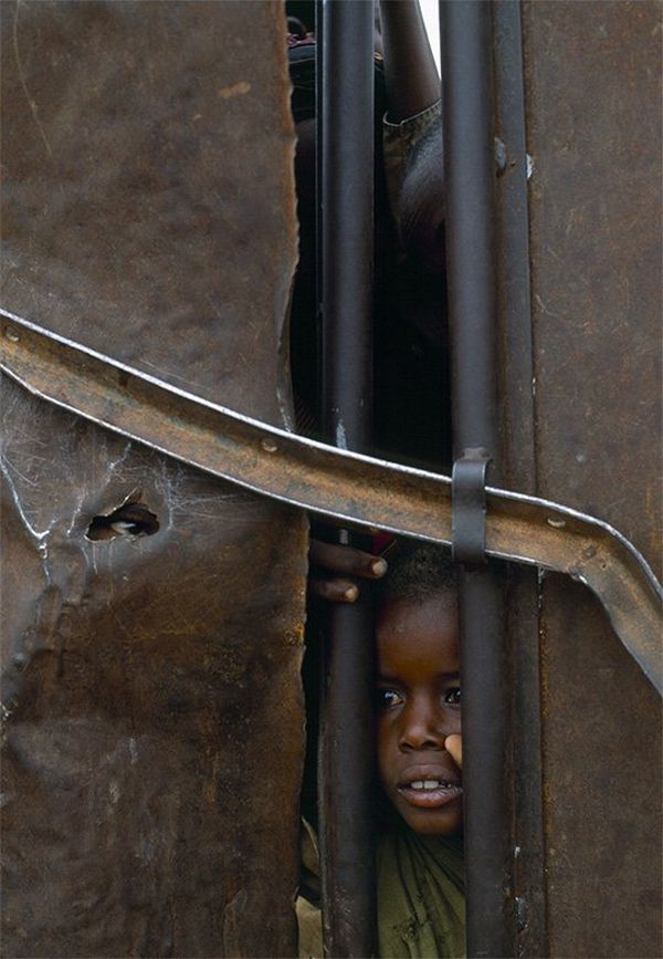 A small child looks through a gap between iron gates.