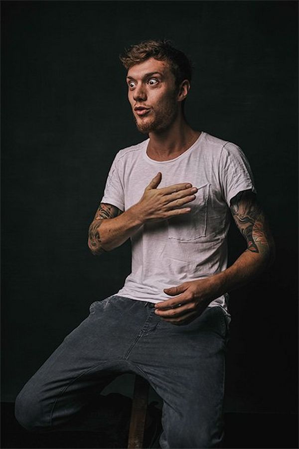 A wide-eyed man with tattooed arms in a white t-shirt.