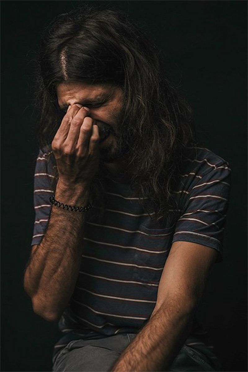 A long-haired, bearded man pinches the bridge of his nose with one hand.
