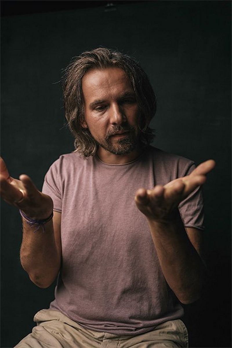 A long-haired man with close-trimmed beard holds both hands up in front of himself, palms upwards.