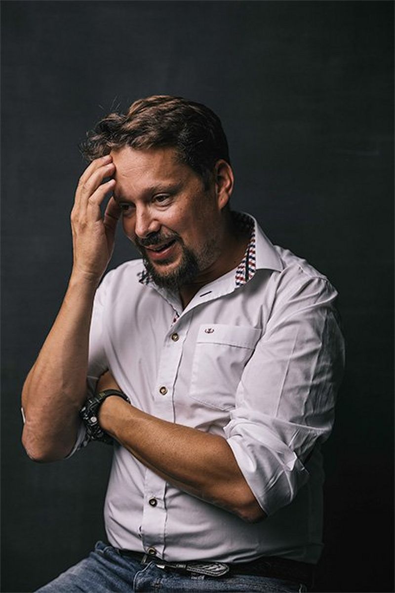 A man with short hair and close-trimmed beard crosses one arm across his body and holds the other up to his forehead.