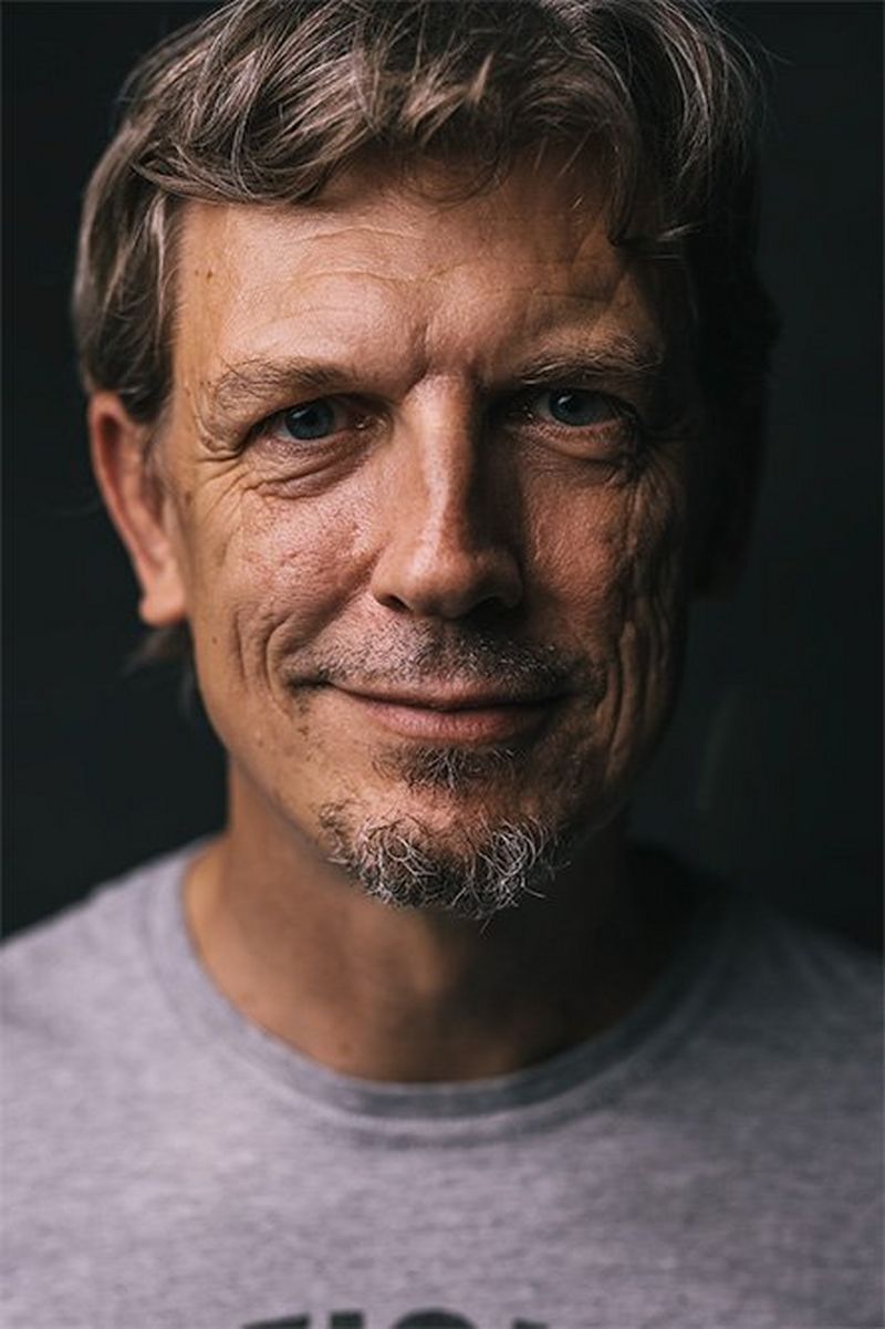 A close-up of a man with greying facial hair, wearing a t-shirt, smiling slightly.