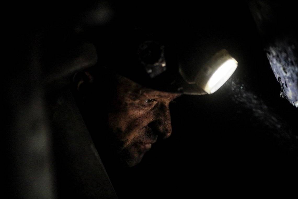 A close-up of a miner’s face looking down, lit by his headlamp 
