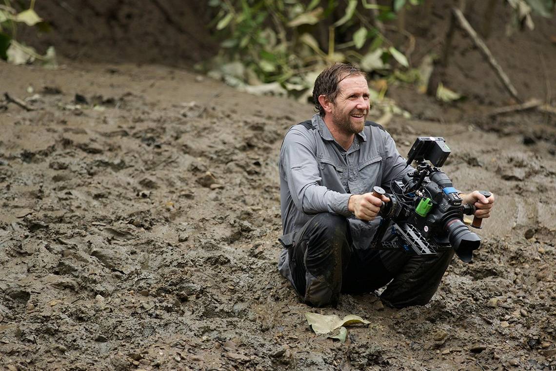 A rain-soaked but smiling Danny sits in several inches of mud holding a camera in both hands.
