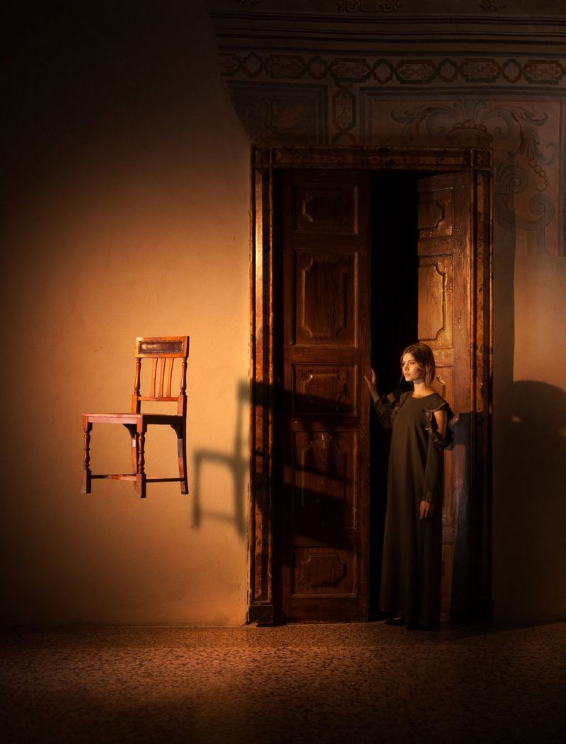A woman stands by a wooden door, while a chair appears to be suspended in the air. Photo by Eberhard Schuy