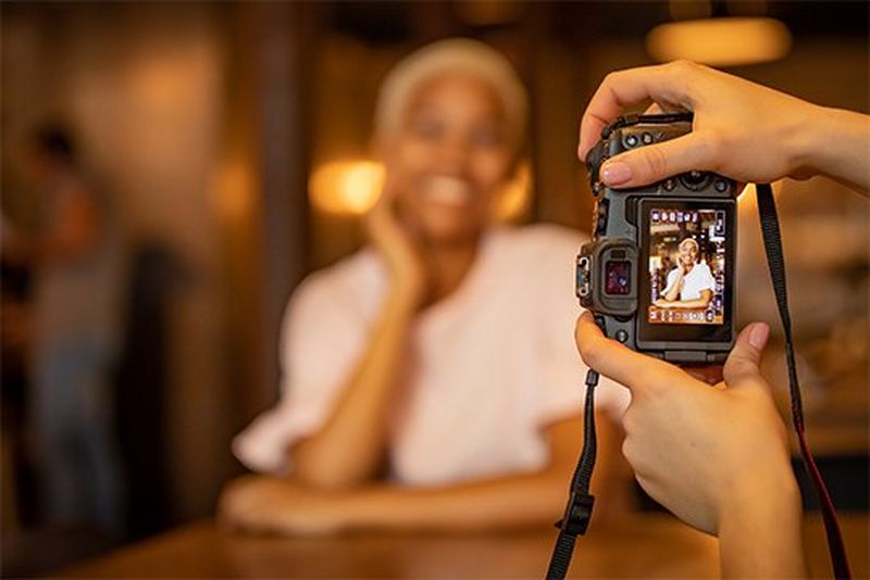 A photographer's hands hold a Canon EOS RP as she photographs a smiling woman in a dimly-lit interior.