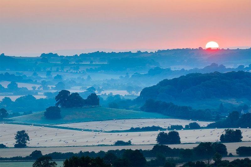 The sun rising over a hilly landscape, with fields of crops in the foreground. Photograph by David Noton.