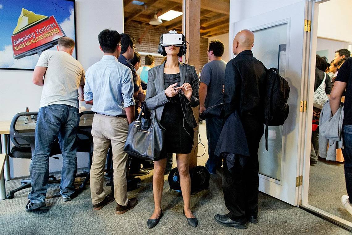 A well-dressed young woman with a large handbag stands facing the camera wearing a VR headset and earphones, completely absorbed, while everyone else in the room is facing the opposite direction.
