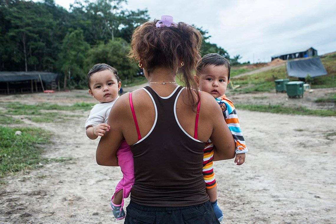 A female ex-member of Columbian militant group FARC stands with her back to the camera, holding two infant children in her arms who look into the camera.