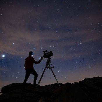 Silhouetted against the night sky, an astrophotographer stands on steep terrain with a Canon EOS Ra attached to a telescope.