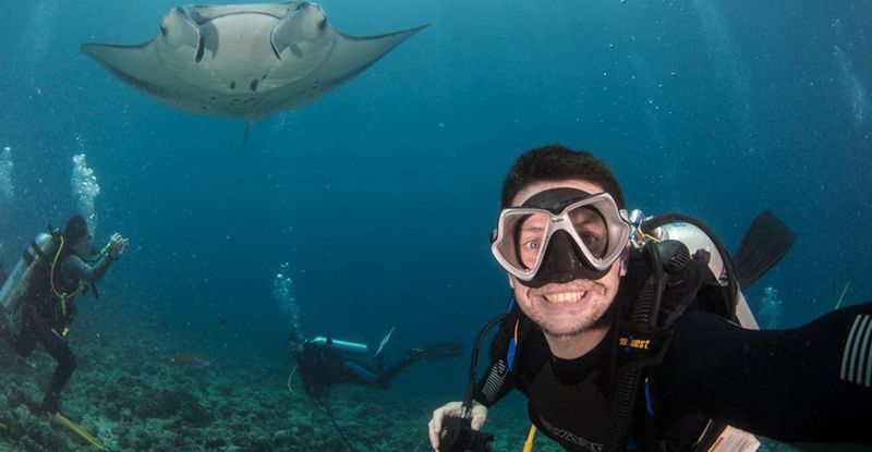 Fergus Kennedy is underwater in goggles and diving gear, though he has removed his breathing mask to smile for the camera. Behind him is a manta ray and two other divers.