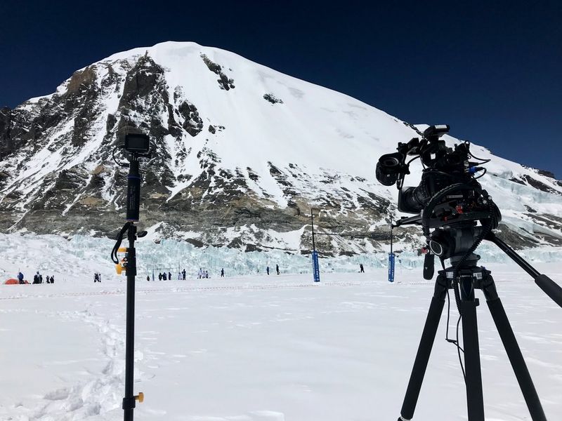 A Canon EOS C200 camera set up on a tripod to film a rugby match in the shadow of a snow-covered mountain.