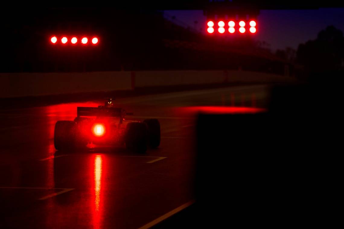 A low-light photo from the rear of the racing car driven by Fernando Alonso, its brake lights emitting a red glow on the wet track.