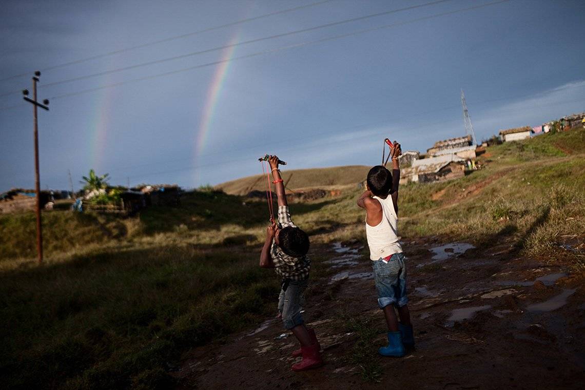 Two boys direct their slingshots to a sky filled with a double rainbow and telephone cables.
