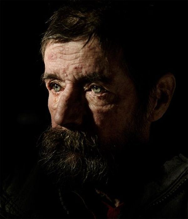 A man with wrinkled skin, a beard and green eyes cries. The picture shows just his face, and is dimly lit.