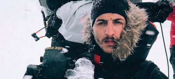 Cinematographer Giacomo Frittelli, wearing a dark hooded parka, holds a video camera in the snow.