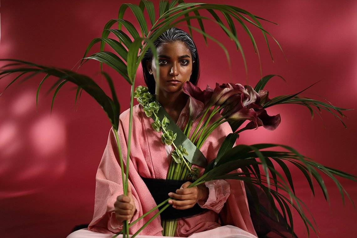 An Indian model holds palm leaves and calla lilies that artfully sit around her, against a deep red backdrop in a studio portrait. 