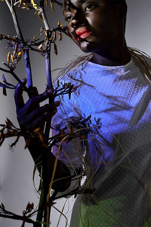 A dark portrait of a black model with coloured blue and green light projected onto her as she holds a sculptural branch.