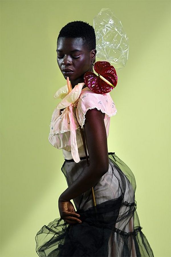A black model poses against a light green backdrop with pink and black outfit and a dark red flower arranged on her shoulder. 