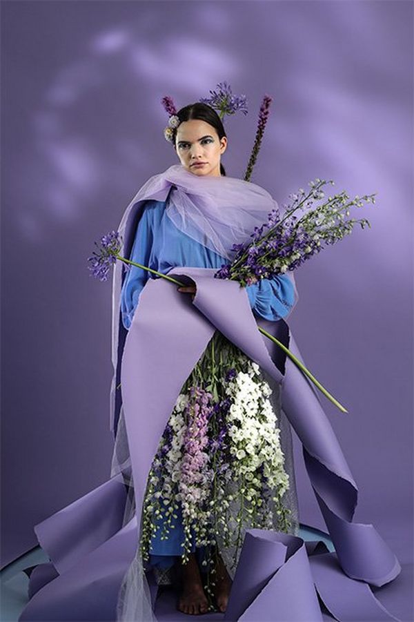 A white model with dark hair holds a lot of flowers and swathes of purple torn paper. She stands against a purple backdrop with dappled light projected onto it.