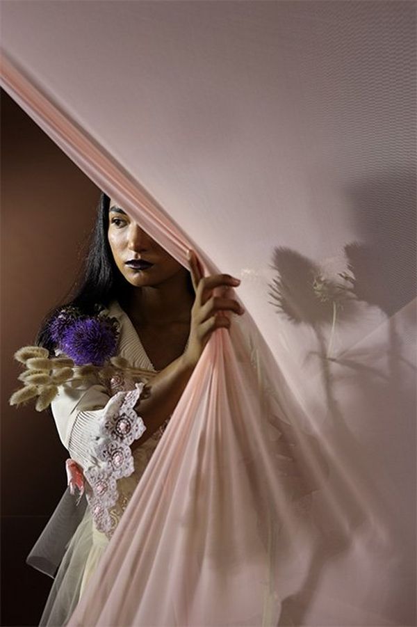 An Indian model pulls a light pink partition to the side, revealing herself standing behind. 