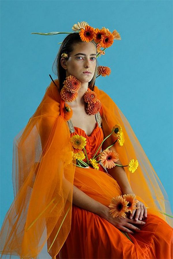 A striking sitting portrait of a white model with intense orange fabric and flowers arranged over her, against a cyan backdrop.
