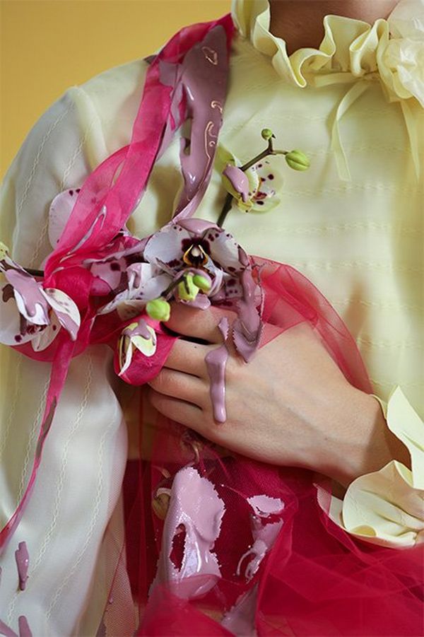A close-up photograph of a white models hand holding orchids, fabric and with paint dripping off her shoulder and hand.