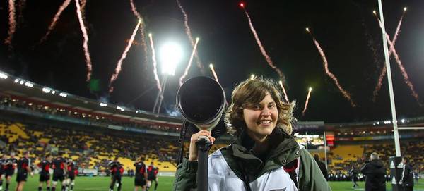 Photographer Hannah Peters holds a Canon camera as she stands in a rugby stadium, fireworks in the background.